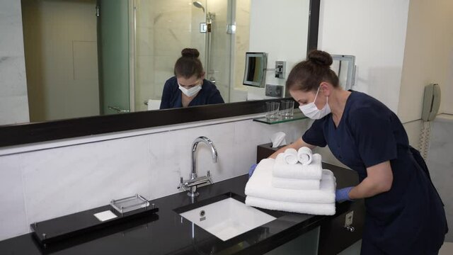 Masked housemaid in uniform and gloves bringing stack of clean, washed towels to hotel bathroom. Woman hotel worker serving bathroom before guest's arrival
