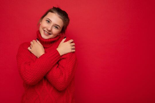Attractive cute smiling young woman wearing warm red sweater isolated over red background wall keeping hands on chest looking at camera