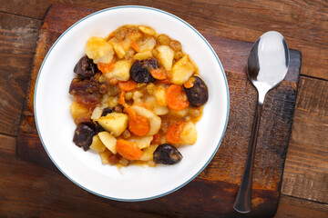 A dish of Jewish cuisine tsimes with carrots and dates in a white plate on a wooden board next to a spoon.
