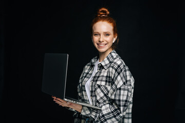 Happy charming young business woman or student holding laptop computer and looking at camera on isolated black background. Pretty redhead lady model emotionally showing facial expressions, copy space.