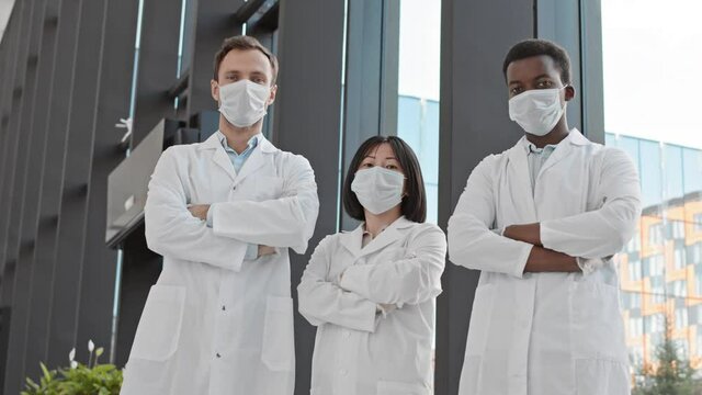 Panning of two diverse male doctors and their female Asian coworker wearing white medical coats standing in modern hospital hall, crossing arms on their chests while looking at camera