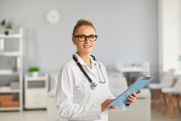 Profile staff. Portrait of smiling female doctor with glasses, therapist, general practitioner looking at camera, standing in hospital with clipboard in hands. Concept of professional doctors.