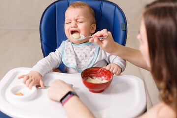Mother tries to feed with spoon naughty infant kid refusing eating - obrazy, fototapety, plakaty
