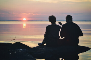 Guy and  girl in love are sitting on the shore of the lake at sunset and sing a song under the...
