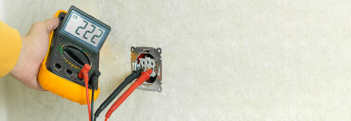 Male electrician checks the voltage of an outlet on a white wall with a multimeter in the house...