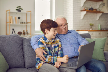 Happy old grandfather and little cute grandson having fun at home using laptop sitting on sofa. Boy teaches his grandfather to use social networks. Leisure concept for two generations.