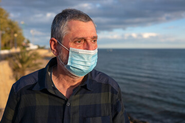 An elderly man stands sad on the beach and looks at the sea at sunset. He has a surgical mask and a tear under his eye. The senior looks unhappy.