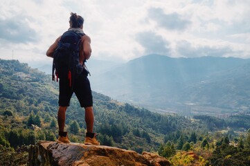 Young man standing with long hair without shirt and big backpack on rock in the mountain looking the city