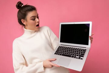 Photo of beautiful amazed and surprised woman with gathered brunette hair wearing white sweater holding computer laptop and looking at the open netbook isolated over pink wall background