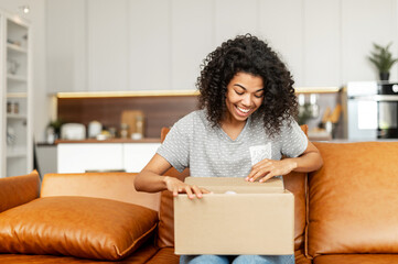 Positive young African American woman sitting on the couch with carton box on laps, feeling curious about ordered item from an online store, smiling client satisfied with fast courier delivery service - Powered by Adobe
