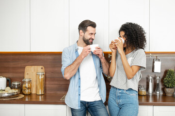 Shy young African American woman and handsome man interracial couple enjoying the happy morning together, standing leaning on the modern kitchen counter and holding cup of coffee in their hands - Powered by Adobe