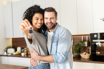Young interracial married couple smiling cheerfully and showing keys from a new apartment, hugging...