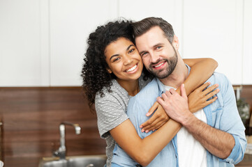Close-up of happy interracial couple posing over blurred kitchen background, happy owners of new flat smiling and looking at the camera, young African American woman hugging handsome man from behind