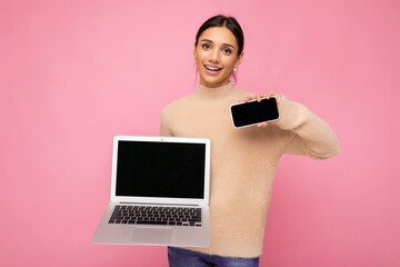 Photo of charming pretty beautiful smiling young woman with dark hair looking at camera holding computer laptop and mobile phone with empty monitor screen with mock up and copy space wearing light