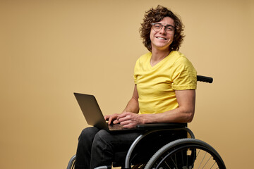 disabled man sits in a wheelchair with laptop, working online. curly young male in casual wear smiles at camera isolated in studio on beige background