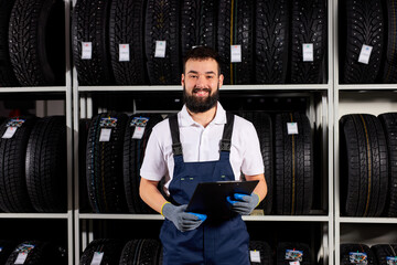 male mechanic with clipboard near tires in automobile service center looking at camera and smiling, in blue uniform