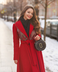 Long-haired woman in red maxi coat and stands near exquisite building