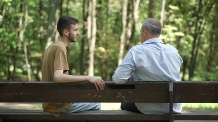 Back view of relaxed young and middle aged Caucasian men sitting on bench in summer park talking. Carefree son and father enjoying sunny day outdoors. Family leisure and lifestyle.
