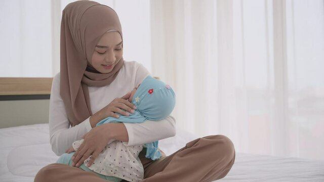 Happy muslim mother holding and kissing adorable baby daughter asleep wearing hijab in her arms on white bed in bedroom. Arab young mom wearing headscarf and sitting on bed while holding her baby. 
