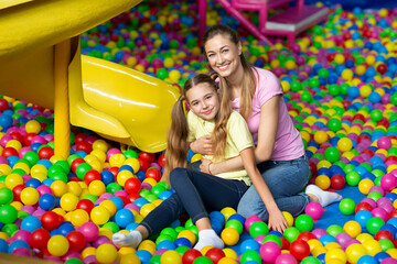 Joyful young mother and her adolescent daughter hugging in ball pit at indoor children playground,...