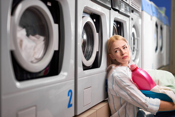 tired woman sitting near washing machines and looking up sadly. Caucasian woman spent time on...