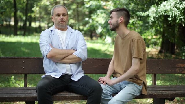 Cheerful Caucasian adult son apologizing as middle aged father sitting on bench with crossed hands. Argued family in sunny summer park outdoors talking. Conflict and lifestyle.