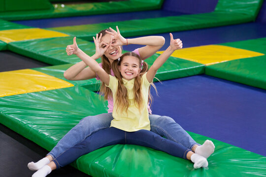 Playful silly mom with her teen daughter sitting at trampoline area of entertainment centre, showing thumbs up gesture
