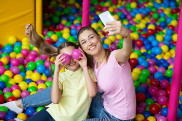 Beautiful young woman with her teen daughter taking selfie together at ball pond in kids amusement centre