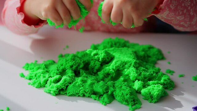 Close up of children's hands playing with green kinetic sand on white table; selective focus