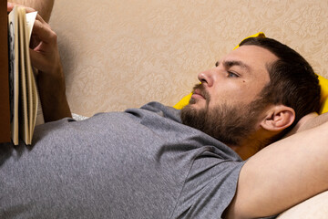The adult man lays on sofa and reads book. A brutal man passionate about reading. Close-up...
