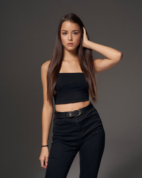 studio portrait of young slim tanned caucasian girl in black jeans and bando top standing and posing against grey studio background