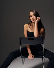 studio portrait of young slim tanned caucasian girl in black jeans and bando top sitting at chair and posing against grey studio background