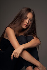 Full length studio portrait of young slim tanned caucasian girl in black jeans and bando top sitting and posing against grey studio background