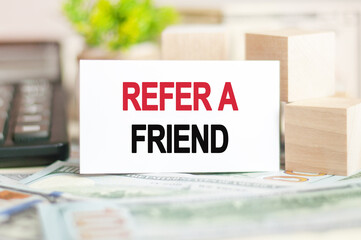 REFER A FRIEND text on white paper, on the background of bills and wooden blocks, business concept
