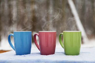 Three cups of coffee, tea on a snowy background, a winter forest. Blue, pink, light green cups on the background of a winter forest landscape.