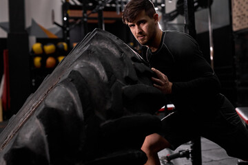 Fototapeta na wymiar Confident male athlete doing tire-flip exercise indoors in modern gym. Cross fit and workout. Strong and handsome athlete in sportswear. sport, healthy lifestyle