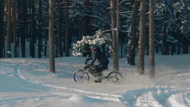 Cyclist in Black Clothes, Helmet and Snow Glasses Riding on Electric Bike in the Snow in Winter Forest. E-Bike Riding and Drifting at Cold Sunny Day - Slow Motion