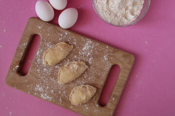 Raw cooking dumplings lie on a board beside egg flour on a pink background view from above