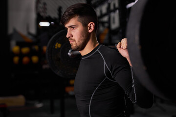 Fototapeta na wymiar Sportsman exercising with barbell. Young caucasian muscular male bodybuilder doing weight lifting workout at dark gym, using sportive equipment, man stand looking at side, side view