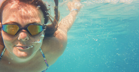 Young woman in reflective swimming glasses swim under water in sea, sun lit bubbles over her face, empty space right side