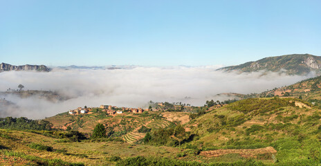 Fototapeta na wymiar Fog over valley with small village on sunny morning in region near Alakamisy Ambohimaha, Madagascar