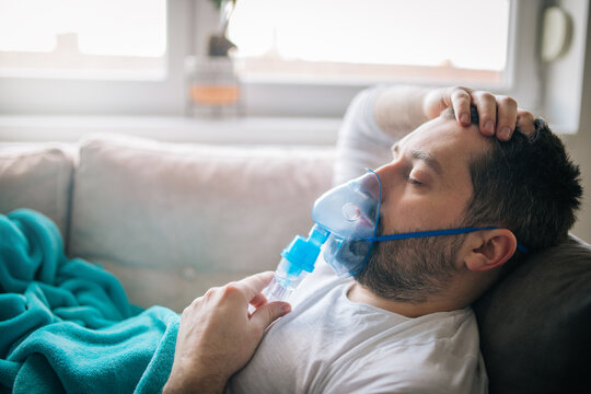 Mature adult man lying down on sofa in living room and using inhaler.He is wearing sleepwear