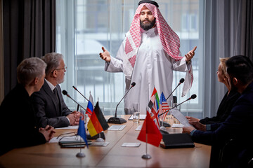 Arab Sheikh holds a meeting without ties to discuss ideas and issues on the agenda, using microphone for giving speech. in boardroom, multi-ethnic colleagues gathered together