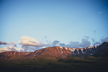 Atmospheric mountain scenery with yellow clouds in blue sky. Scenic landscape with illuminating sunset clouds above mountains with snow. Beautiful sunrise in mountains. Snow on rocks in sunlight.
