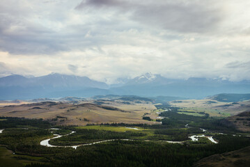 Vintage landscape with vast plateau with mountain river and forest on background of snowy mountain ridge under cloudy sky. Green mountain valley and snow mountain range among low clouds on horizon.