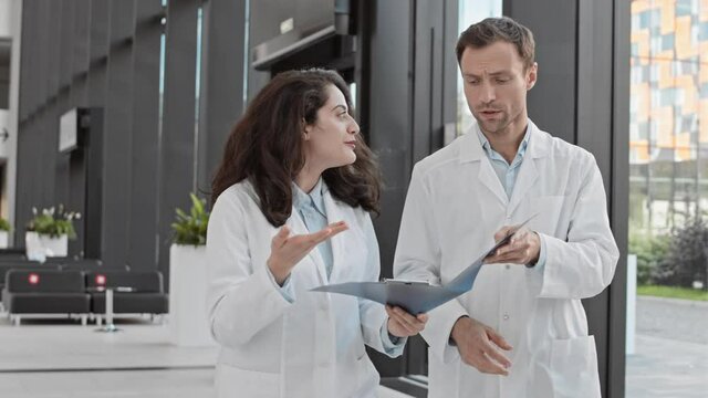 Medium shot of mixed-race female doctor and her male Caucasian coworker going along hospital hall, examining patient card and then looking at camera