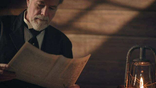 Overhead window light and an oil lantern illuminates this early 19th century scene of a gentleman standing next to a rustic table thoughtfully reading a newspaper.