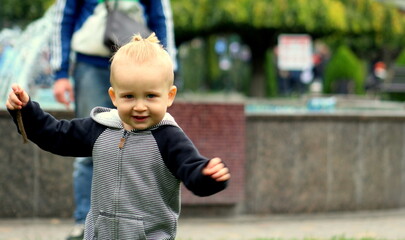 A cute toddler boy blond in a gray overalls stands on the grass in the park.
