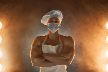 New normal concept. Muscular chef wearing protective medical mask, posing with folded arms on smoky...