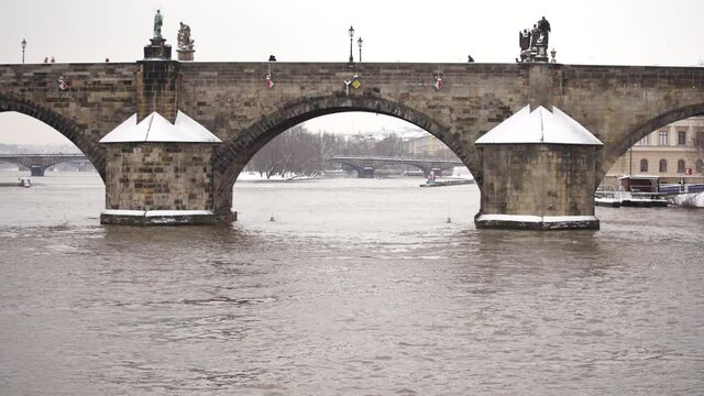 detailed view of the stone on the snowy Charles bridge on the flowing river wavy and snowy statues and street lights on the bridge and silhouettes of pedestrians walking on the bridge in prague 